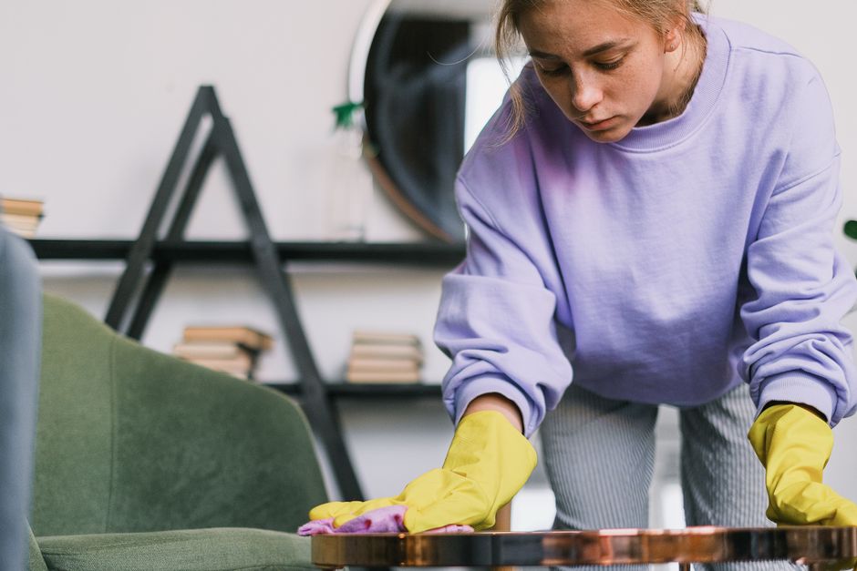 Crop calm female in casual clothes and sterile gloves wiping dusk on wooden table in light room in daytime