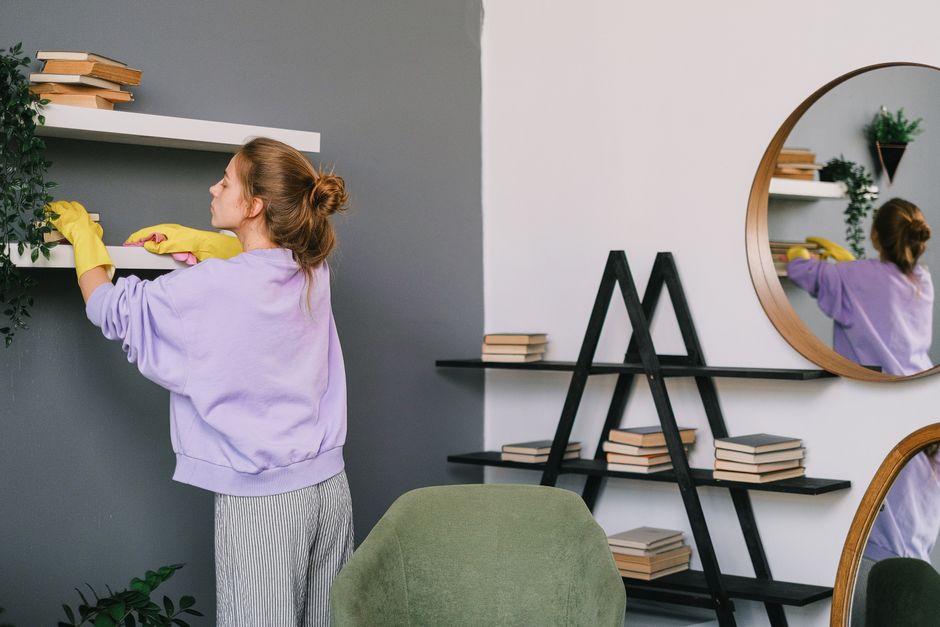 A woman cleans shelves in a stylish living room with mirrors and decor.